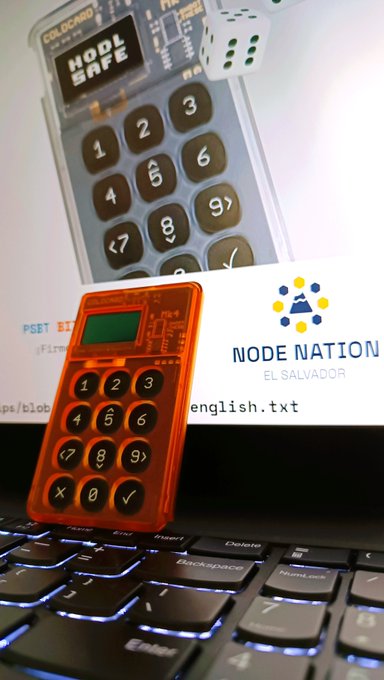 A close-up of a physical crypto hardware wallet (orange keypad device) resting on a laptop keyboard, with packaging labeled 'HODLR SAFE' and a 'NODE NATION EL SALVADOR' card in the background, along with a white dice.