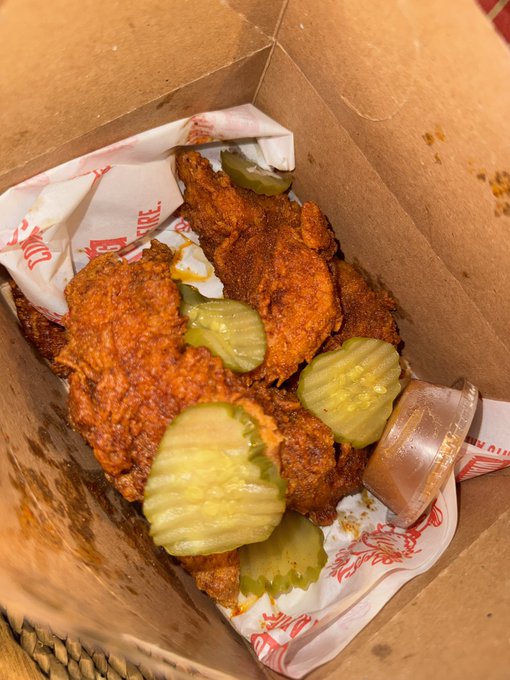 A cardboard box containing several pieces of fried chicken topped with pickle slices, accompanied by a small container of dipping sauce.