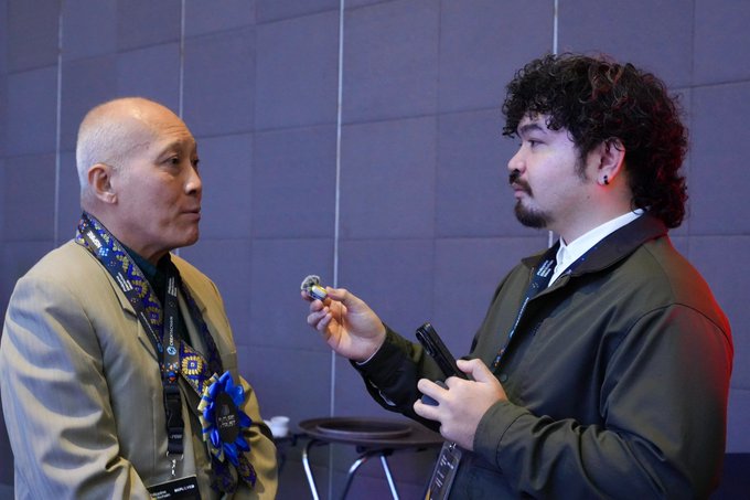 The image shows a man wearing a suit and tie holding up an object while talking to another person who is also dressed in formal attire. The man with the object appears to be presenting something or explaining its purpose, possibly related to tokenization or fintechs. The other person seems interested in what the first man has to say.