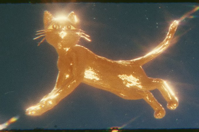 A golden, glowing cat figurine or decoration shown against a dark blue background with film grain effects.