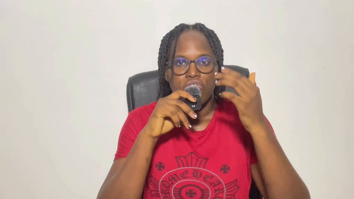 The image features a man wearing glasses, sitting at a desk with his hands on his face while holding a microphone. The background of the image is white, which contrasts with the red shirt he's wearing. There are no other objects or people visible in the photo.