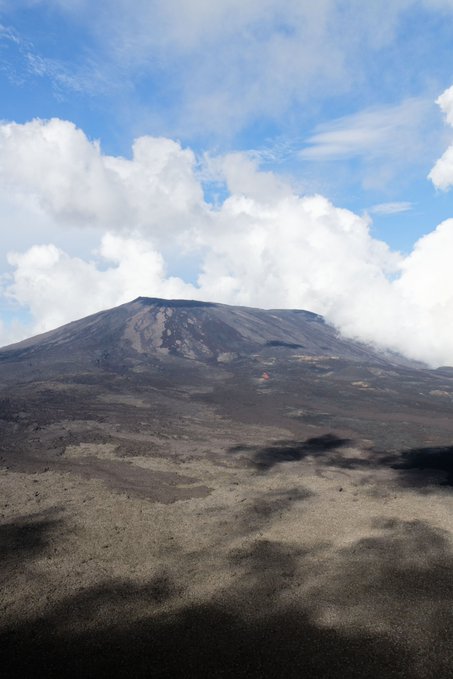 A volcanic mountain rises above a barren, rocky landscape under a blue sky filled with fluffy white clouds.