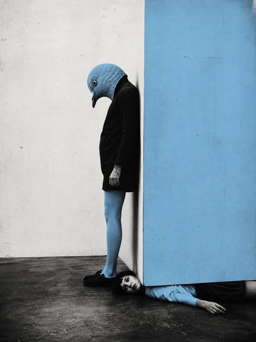 A surreal artistic photograph shows a person wearing a large blue bird mask and black shirt standing against a wall, with another person's head and arm visible emerging from underneath the wall panel.