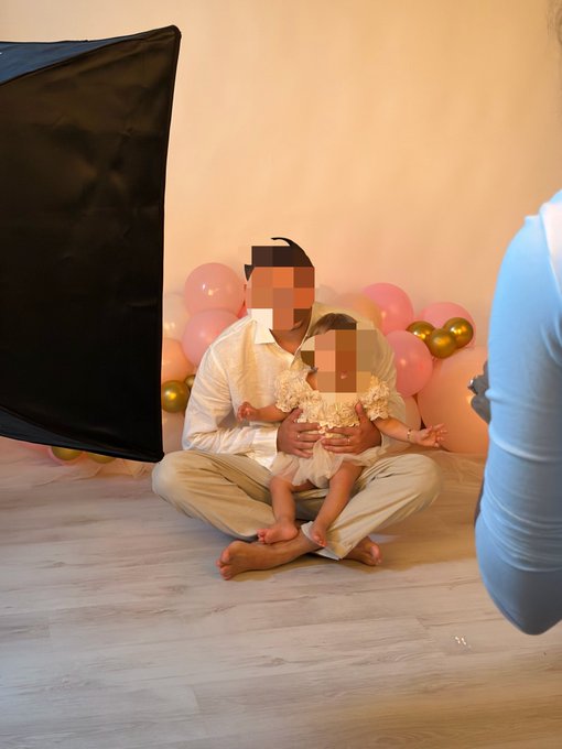 A behind-the-scenes photo from a photoshoot showing a father and young child (faces blurred) sitting in front of pink and gold balloons, with a black reflector screen to the left