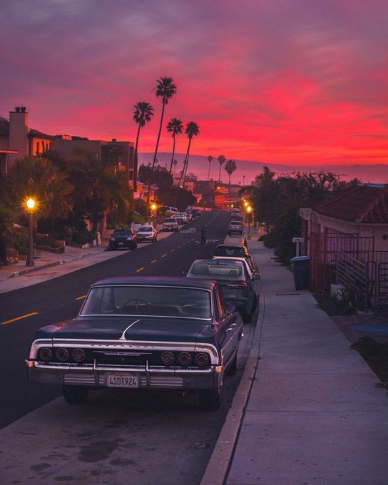 A vintage dark-colored classic car parked on a street with palm trees silhouetted against a vivid pink and red sunset sky.