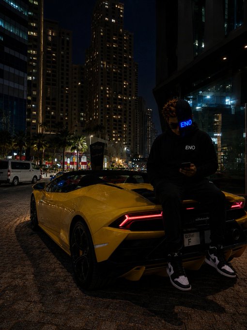 A person wearing an 'ENO' hoodie and a face covering with 'ENO' written on it sits on the back of a yellow Lamborghini Huracán at night in a city, looking at their phone.