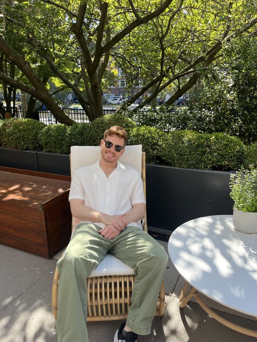 A smiling young man with red hair and sunglasses relaxes in a wicker lounge chair on a sunny outdoor patio, playfully campaigning for a Solana Breakpoint ticket with self-deprecating charm and over-the-top enthusiasm about Solana becoming the "global currency of the universe."