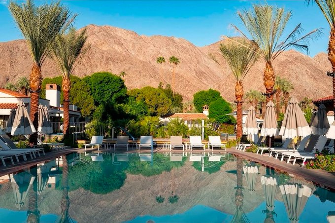 A luxurious resort pool with crystal clear water reflecting palm trees, lounge chairs with umbrellas, and a backdrop of desert mountains under a clear blue sky.
