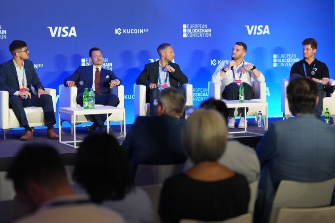 A panel discussion at the European Blockchain Convention featuring speakers seated on white chairs with a Visa and KuCoin-sponsored backdrop, while audience members watch from the foreground.