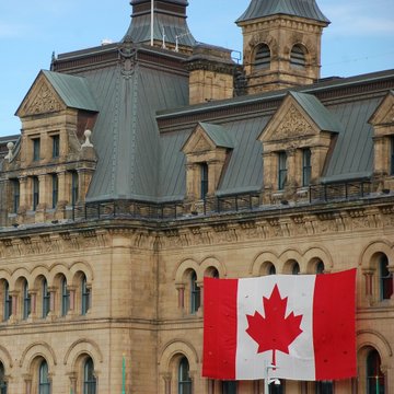 A historic stone building with ornate architecture and copper-green rooftops, with a large Canadian flag hanging prominently in the foreground.