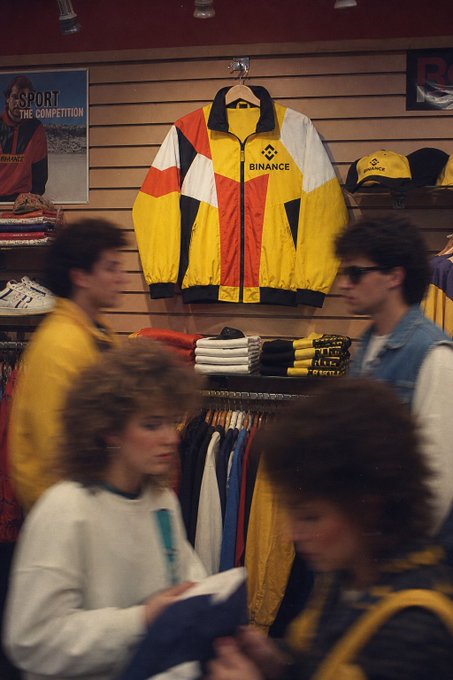 A vintage 1990s-style clothing store displays a bold color-blocked yellow, orange, white, and black jacket with "BINANCE" prominently featured on the chest, alongside matching caps and folded tees, capturing the crypto exchange's branding through retro sportswear aesthetics. Shoppers browse racks of athletic apparel in the foreground while the distinctive Binance merchandise hangs as the centerpiece.