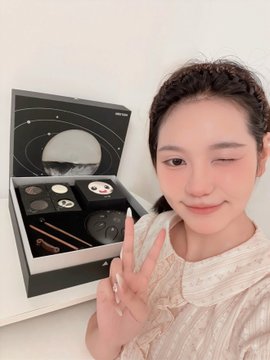 A young woman winking and making a peace sign next to an open makeup gift box containing brushes and cosmetic products.