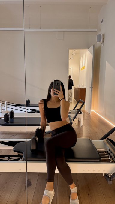 A mirror selfie of a woman wearing a black sports bra and black leggings, sitting on a Pilates reformer machine in a studio with white walls and wooden floors.