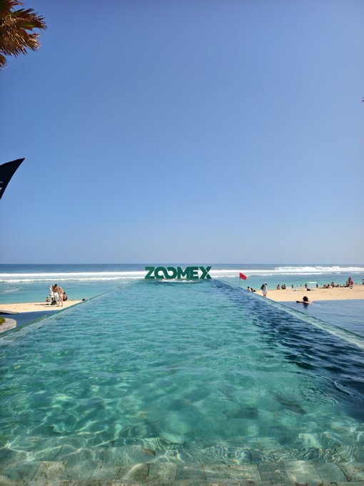An infinity pool overlooking a beach and ocean, with green 'Z.COMEX' letters placed at the edge of the water.