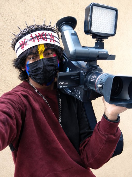 A person wearing a spiked crown headband, black face mask, and maroon sweatshirt holds a large professional video camera (GH series by Panasonic/DJI) while taking a selfie.