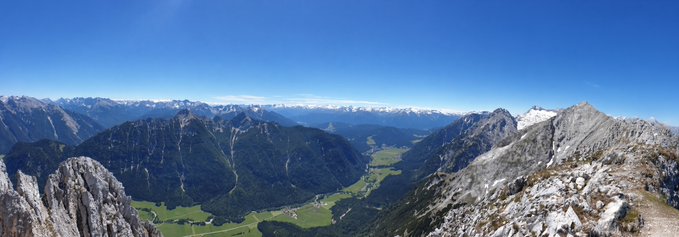 A panoramic mountain landscape showing a valley with green fields surrounded by rocky, forested mountains under a clear blue sky.