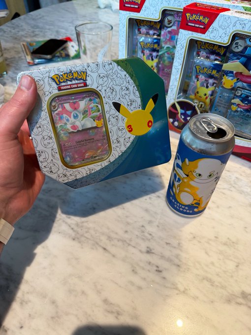 A hand holds a Pokémon Trading Card Game Sylveon VMAX tin next to a Pokémon-themed beverage can on a marble countertop.