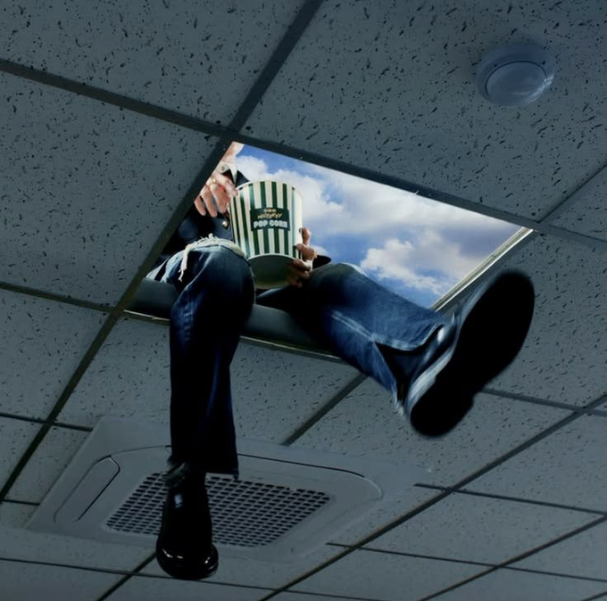 A surreal composite image showing a person sitting on a dropped ceiling tile eating popcorn, with their legs dangling down through the tile into what appears to be an office room with fluorescent lighting.