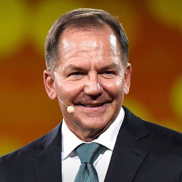 A close-up portrait of a smiling middle-aged man with short hair wearing a black suit and green tie, speaking into a headset microphone against a blurred yellow background.