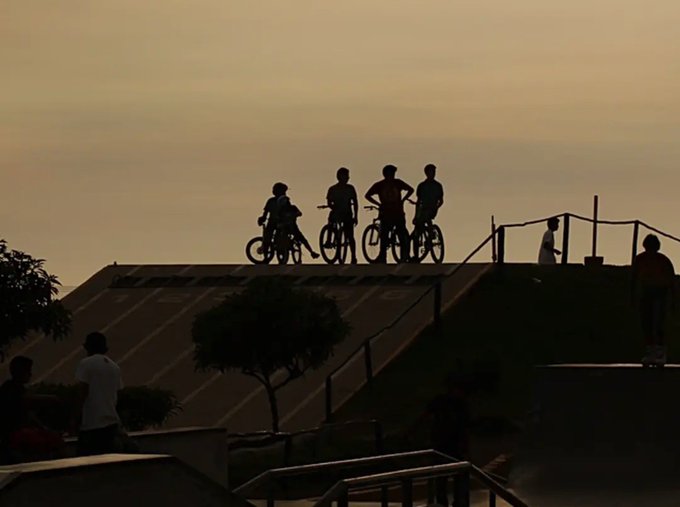 A silhouette of four young people with bicycles stands atop a skate ramp against a hazy golden sky at sunset.