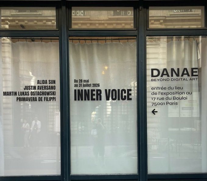 A photograph of a storefront window with white curtains displaying text for the 'DANAE BEYOND DIGITAL ART' gallery, advertising the 'INNER VOICE' exhibition from May 28 to July 31, 2026, featuring artists Alida Sun, Justin Aversano, Martin Lukas Ostachowski, and Primavera de Filippi.