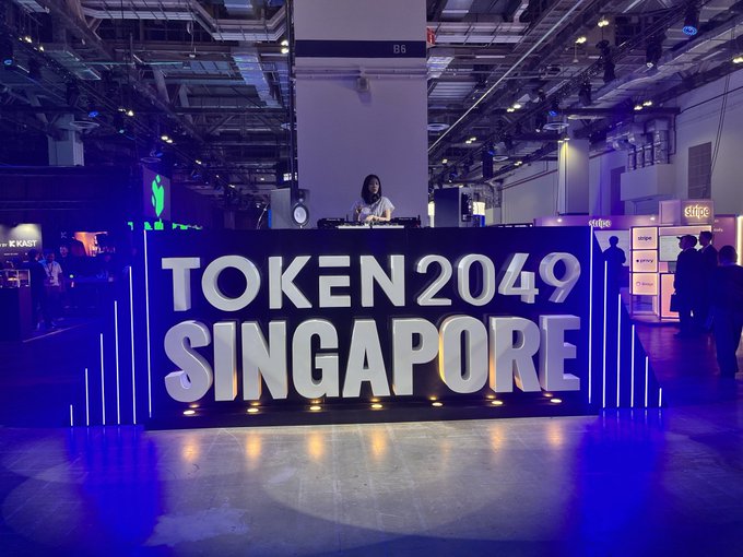 Photo from the TOKEN 2049 SINGAPORE crypto conference showing a DJ booth set up in an expo hall with large illuminated letters spelling the event name and attendees walking around in the background.