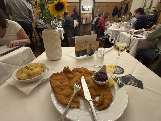 A large schnitzel served with a side of potato salad, cranberry sauce, and a lemon wedge sits on a white table in what appears to be a restaurant, with a glass of white wine and a sunflower in a vase nearby.