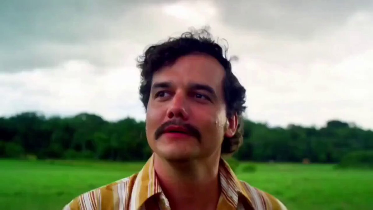 The image features a man with curly hair wearing a striped shirt standing in an open field or park-like setting. The man appears to be looking up at something above him, possibly observing the sky or contemplating his thoughts. The surrounding environment suggests that he is in a peaceful and natural outdoor space.