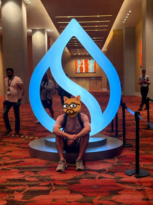 A person sits cross-legged in front of a large blue illuminated water droplet sculpture in a convention center, while another person wears a humorous orange cat face filter on their head.