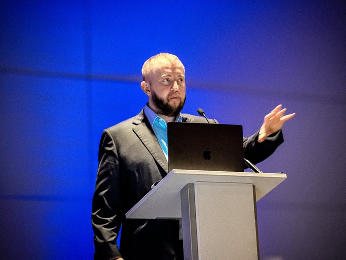 A bearded man in a dark suit and blue tie speaks at a podium with an Apple laptop against a blue backdrop, gesturing with his arm raised.