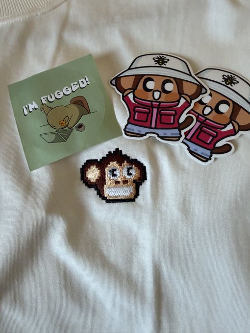 A sticker reading "I'M FUGGED!" featuring a cartoon character sits on a white fabric alongside two character stickers and a pixel-art monkey patch, representing memorabilia from the Chimpers x Fugz meetup.