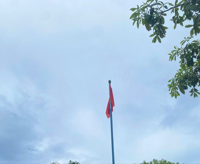 A red flag flies on a tall pole against a partly cloudy sky, framed by green tree branches in the upper right corner. The flag's raised position symbolizes motivation and forward momentum, visually echoing the message's encouragement to push harder and persist for good things to come.