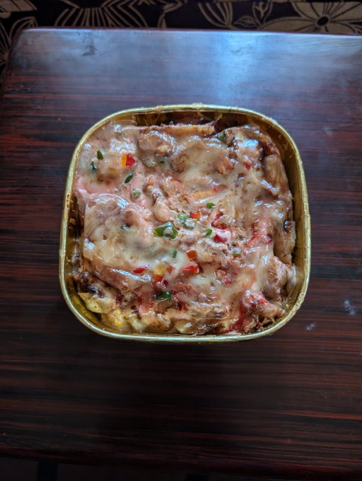 A baked pasta dish topped with melted cheese and colorful peppers sits in a gold takeout container on a wooden table, matching the post's mention of enjoying a favorite pastry-like comfort food during a busy weekend.