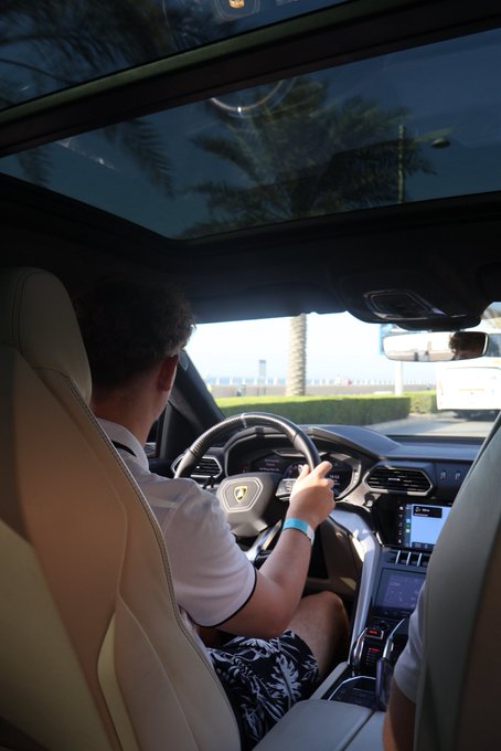 A man drives a luxury vehicle with a Lamborghini emblem on the steering wheel, wearing patterned swim trunks as palm trees pass by through the windshield. The caption "GM (get money)" reflects the aspirational lifestyle depicted, emphasizing the wealth and success associated with high-end cars and tropical settings.