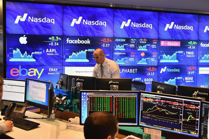 Traders work at the Nasdaq exchange floor beneath large displays showing real-time stock quotes for Apple (AAPL +0.3%), Facebook (-0.5%), Amazon (AMZN +0.1%), Cisco (CSCO +1.6%), T-Mobile (TMUS +0.4%), eBay (EBAY), Alphabet (GOOG -0.5%), and the Nasdaq Composite (NDX -0.27%), illustrating the very U.S. equity markets that are now pushing to extend trading hours to 23 hours—a move aimed at letting Asian investors participate during hours once exclusive to crypto markets.