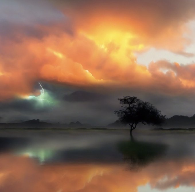 A dramatic landscape photograph shows a lone tree silhouetted against a vibrant orange and dark cloudy sky with a visible lightning bolt, reflected in calm water below.