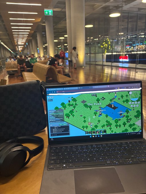 A laptop on a wooden table in what appears to be an airport or large public space displays a voxel-based game map labeled 'The Pond', with black over-ear headphones resting beside it.