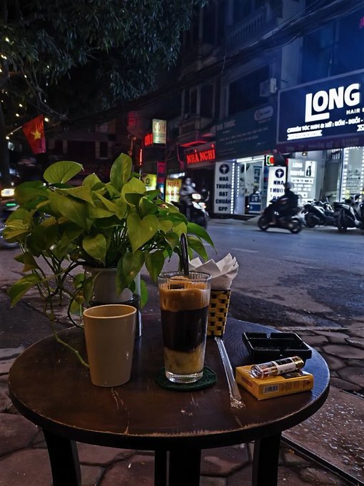 A nighttime street scene in what appears to be Vietnam, with a coffee shop table in the foreground holding a Vietnamese iced coffee, a teacup, and a cigarette pack, with shops like 'LONG' and 'NHÀ NCĐI' visible across the street.