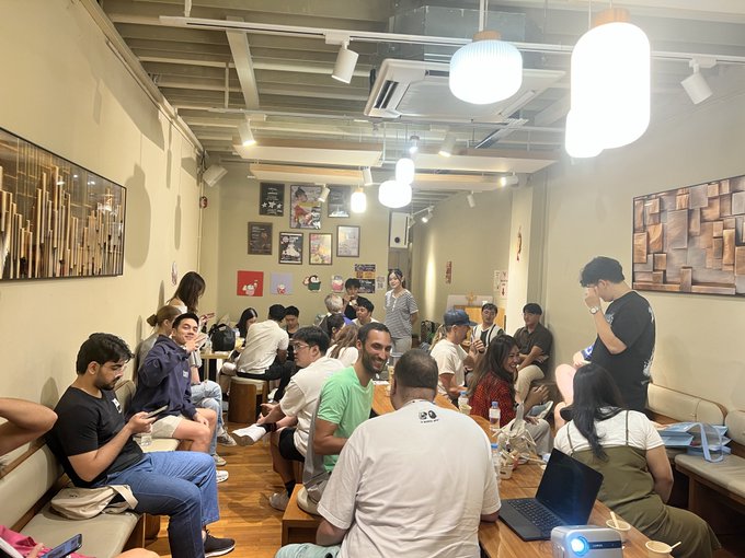 A crowded indoor venue shows a group of people sitting around wooden tables with laptops, illuminated by large hanging pendant lights.