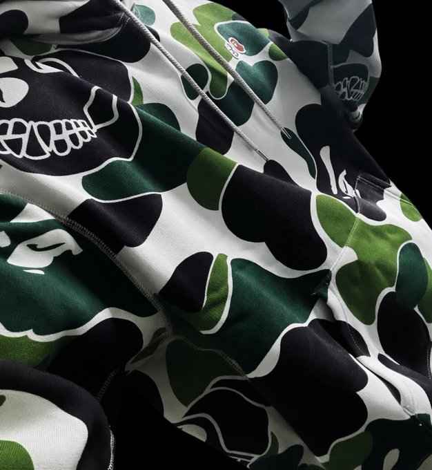 The image features a hoodie with a camouflage pattern, predominantly green and black, with white spots scattered throughout. The hoodie has a zipper running down the front and appears to be made of a soft fabric. The text "BAPE" is visible in the image, which could potentially be related to the brand or style of the hoodie. Additionally, there are two other words that seem unrelated: "Grail" and "Secured". These words might be part of an advertisement or promotional material for the hoodie.