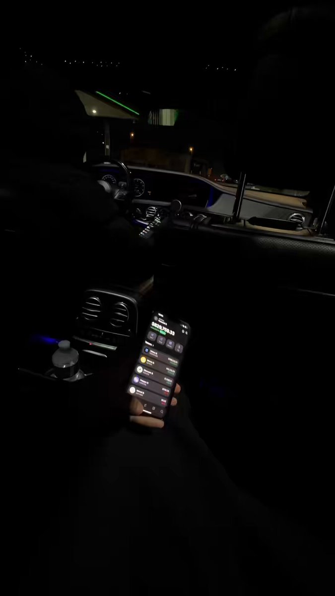 The image shows a person holding up their phone with an app open, possibly for viewing or sharing content. The scene takes place inside a car at night, with the interior illuminated by the glow of the phone's screen. The background appears dark, emphasizing the focus on the phone and its contents.