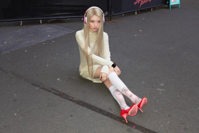 A young woman with long platinum blonde hair and pink headphones sits on the pavement wearing a pale yellow long-sleeve top, white over-knee socks, and bright red stiletto heels.