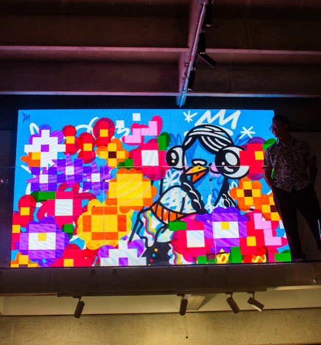 A person standing next to a vibrant, colorful digital artwork displayed on a large screen, featuring a cartoon character surrounded by pixelated flowers and gift boxes.