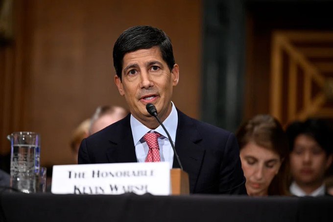 A man in a dark suit with red tie speaks at a hearing microphone, with a nameplate reading 'THE HONORABLE NEIL WARSH' visible.