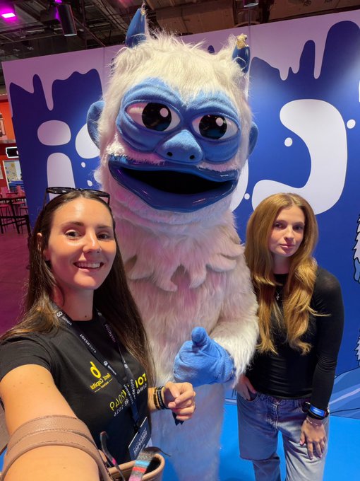 Two women pose for a selfie with a person in a blue and white furry yeti mascot costume at what appears to be a QuBit conference booth.