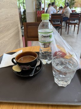 A cafe table setting with a small coffee cup, a bottle of Dasani water, a glass of ice water, and pastries, with people dining in the background.