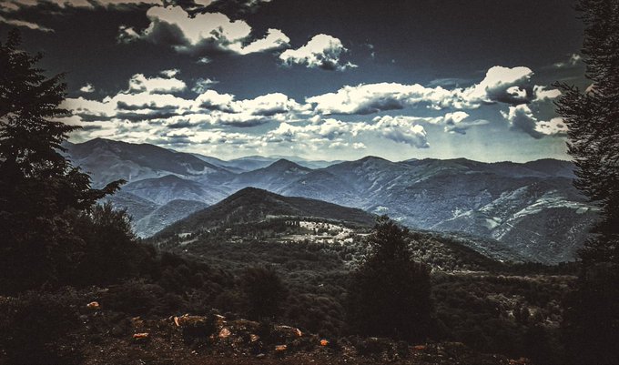 A dramatic landscape view of mountain ranges layered in the distance under a dark sky filled with scattered clouds, framed by dark silhouettes of trees in the foreground.