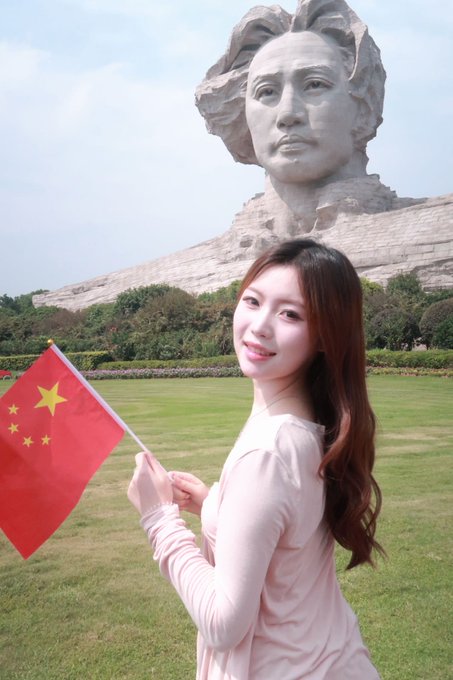 A young woman in a pink dress smiles at the camera while holding a Chinese flag, with a large Mao Zedong sculpture visible in the background.