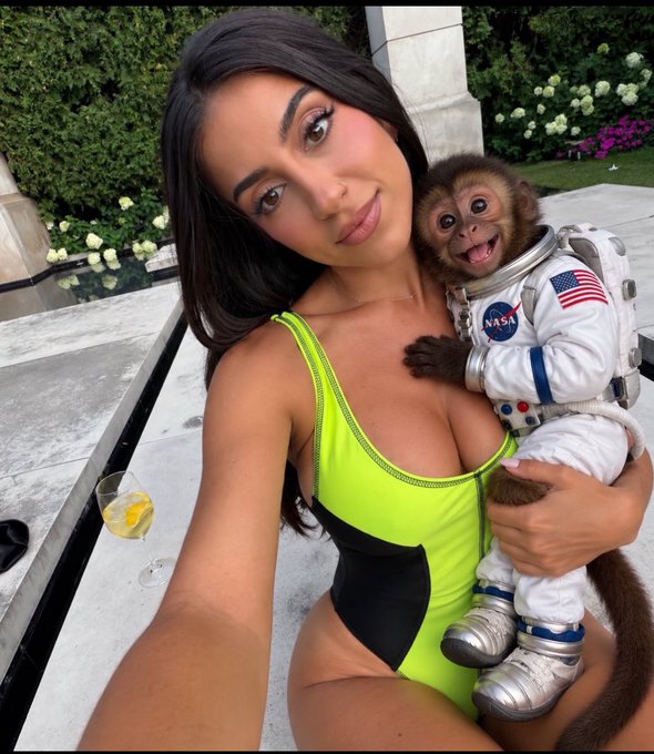 A woman with long dark hair wearing a neon yellow and black swimsuit holds a small monkey dressed in a NASA astronaut suit, taking a selfie outdoors.