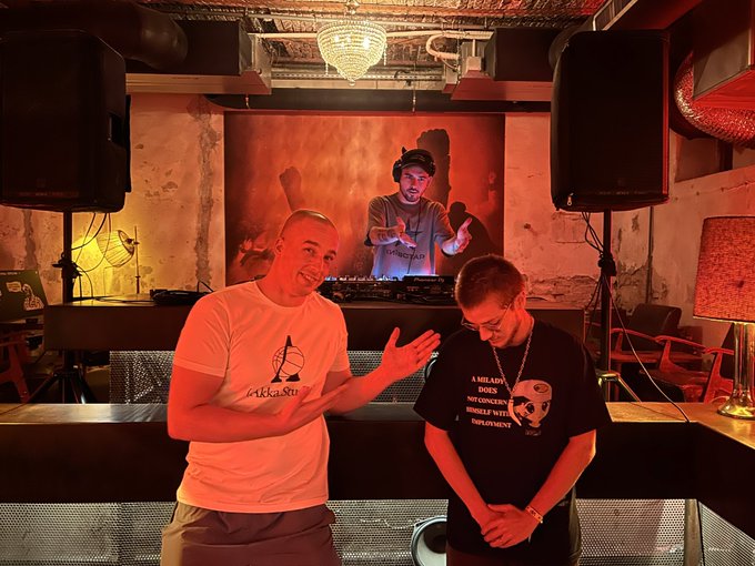 Two men stand in a dimly lit venue with red lighting; one wears a white t-shirt and the other a black graphic t-shirt, with a DJ visible behind them.